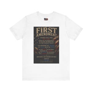 May include: A white short-sleeved t-shirt featuring a central rectangular graphic with a dark background. The graphic displays the text of the First Amendment of the U.S. Constitution overlaid on a faded American flag design in red, white, and blue.