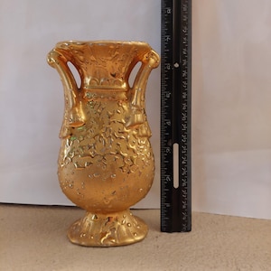 Könnte beinhalten: Eine goldfarbene Keramikvase mit zwei Henkeln und einer strukturierten, gesprenkelten Oberfläche. Die Vase steht neben einem schwarzen Lineal, das eine Höhe von etwa 19 cm anzeigt. Das Gefäß hat einen breiten Rand und einen Standfuß.