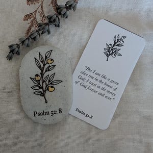 May include: A speckled oval stone featuring a black line drawing of an olive branch with yellow accents and the text Psalm 52:8, alongside a white rectangular card printed with the same olive branch and the full text of Psalm 52:8.