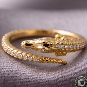 Può includere: Un anello dorato a forma di coccodrillo che si avvolge attorno al dito. La testa e il corpo del coccodrillo presentano una superficie testurizzata a scaglie con piccole pietre rotonde trasparenti incastonate lungo il dorso. Il testo KHODALGLITZ è visibile.