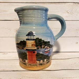 May include: A ceramic pitcher with a light blue glazed finish featuring a hand-painted scene of a lighthouse, green trees, and a small sailboat on the water. The vessel has a curved handle, a wide rim, and a tan base with visible brush strokes.