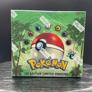 May include: A sealed green Pokémon trading card game booster box featuring a central red and white Poké Ball graphic, various elemental energy symbols, and the text Pokémon 1st Edition Limited Printing with Wizards of the Coast branding.