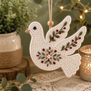 May include: A cream-colored crocheted dove ornament featuring embroidered green leaves and small red berries on its wings and body. The bird has a single black bead eye, a light brown beak, and is suspended by a tan string with a small wooden bead.