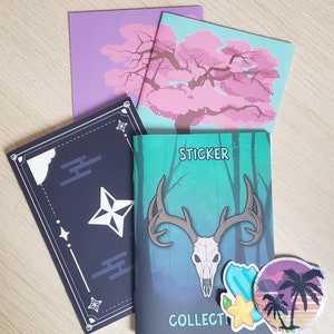 Può includere: Tre piccoli quaderni con design diversi. Il primo quaderno ha una copertina nera con una stella bianca e motivi geometrici. Il secondo quaderno ha una copertina rosa e blu con un albero rosa. Il terzo quaderno ha una copertina verde con un teschio di cervo e il testo "Sticker Collector".