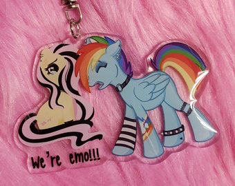 May include: A clear acrylic keychain featuring a star-shaped clasp and two cartoon ponies. One pony is cream-colored with long black hair, and the other is light blue with a rainbow mane and tail. The text "We're emo!!!" is printed in black.