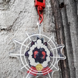 May include: A clear acrylic keychain featuring a black and white cartoon sheep with red eyes and a red bell. The sheep is surrounded by a white spiderweb design.