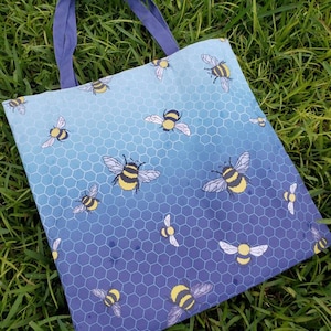 Może przedstawiać: Niebieska i fioletowa torba typu tote z wzorem plastra miodu i pszczołami. Torba ma niebieskie płócienne paski, a na dole nadrukowany jest tekst "Art by Pineathers".