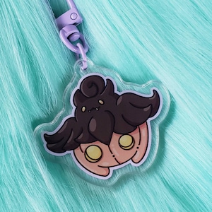 May include: A clear acrylic keychain featuring a cartoon illustration of a pink and brown creature with yellow eyes. The creature has a large, dark brown head with a curled tuft of hair and wings. The keychain has a purple metal clasp.