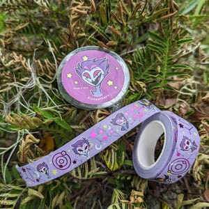 May include: Two rolls of decorative washi tape. One roll is round with a purple and pink design and the text "Pinfeathers". The other roll is unrolled, showing a repeating pattern of a cartoon character, stars, and moons. The tape is 15mm x 10m.