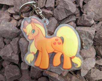 May include: A clear acrylic keychain featuring a cartoon pony with an orange body, yellow mane and tail, and green eyes. The pony has a cutie mark of three red apples on its flank and is attached to a silver-colored metal split ring keyring.