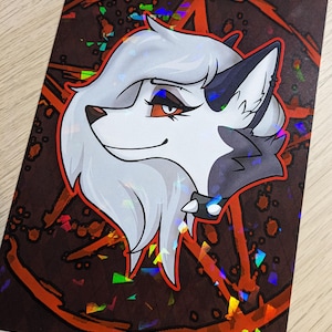 Puede incluir: Un lobo blanco y gris con ojos rojos y una gargantilla negra, con un pentagrama detrás. El fondo es marrón oscuro con acentos rojos y naranjas y un efecto holográfico.