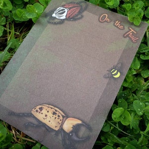 May include: A brown paper notepad with a green and brown watercolor background. The notepad has a title that says "On the Trail" and features illustrations of a beetle, a bee, and a stag beetle.