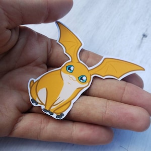 May include: A yellow and white cartoon sticker of a creature with wings and blue eyes. The creature is sitting on its hind legs with its wings spread out.