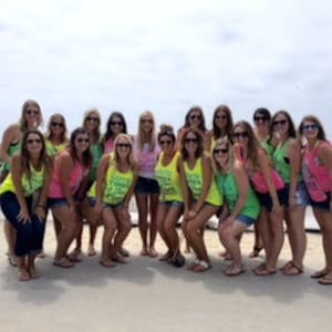 May include: A group of women wearing matching tank tops with the text "Squad Goals" in green and pink letters. They are standing on a beach with the ocean in the background.