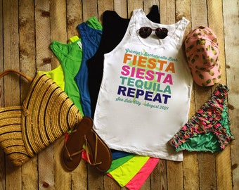 May include: A white tank top with the text "Gricely's Bachelorette Bash Fiesta Siesta Tequila Repeat Sea Isle City - August 2021" printed on it. The tank top is surrounded by other clothing items, including a straw bag, a pink hat with pineapple designs, and a pair of brown sandals.
