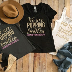 May include: Three shirts for a bachelorette party. One black shirt says "We are popping bottles Heather's Bachelorette". One gray shirt says "We are popping bottles Heather's Bachelorette". One white tank top says "Pop the bubbly I'm getting a hubby".