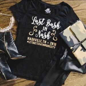 May include: Black t-shirt with gold lettering that says "Last Bash in Nash" with the text "Nashville, TN - 2018 #LetsGetCrayinMusicCity" underneath. The shirt is laying on a wooden surface with black leather pants, black heeled boots, and a camouflage clutch.