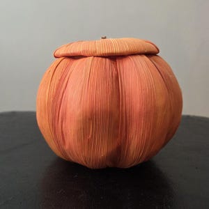 May include: A round, orange decorative object crafted from dried corn husks, featuring a textured, ribbed surface that mimics the appearance of a pumpkin. The object includes a matching lid with a small, central stem-like detail at the top.