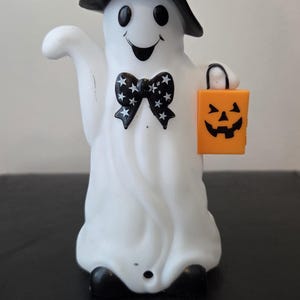 May include: A white ghost figurine wearing a black witch hat and a black bow tie with white stars. The ghost has a smiling face and holds a small orange rectangular trick-or-treat bucket featuring a black jack-o'-lantern face design.