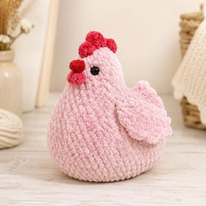 May include: A plush crochet chicken toy made from soft, light pink yarn. The chicken features a bright red textured comb and wattle on its head, along with a single black bead eye. The body is rounded and plump with a small, textured wing.