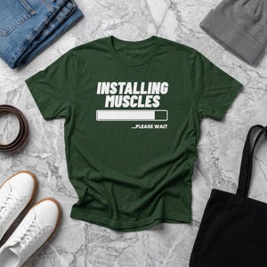 Puede incluir: Una camiseta de manga corta de color verde oscuro con el texto blanco "INSTALLING MUSCLES" sobre un gráfico de barra de carga y el texto "...PLEASE WAIT". La camiseta tiene cuello redondo y está hecha de tela lisa con un diseño estampado.