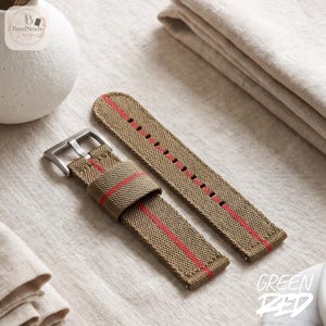 May include: A khaki-colored watch strap made of woven fabric featuring a single vertical red stripe running down the center. The strap includes a silver-toned metal buckle and a matching fabric keeper loop, with pre-punched holes for adjustment.
