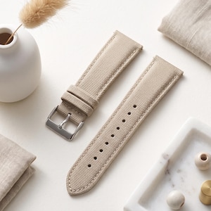 May include: A beige watch strap made of a textured fabric material, featuring a silver-toned metal buckle and two matching fabric keepers. The strap has a series of punched holes for adjustment and visible stitching along the edges.