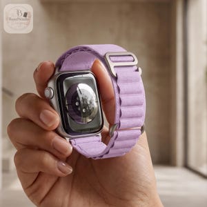 Può includere: Uno smartwatch argentato con un cinturino in tessuto color viola chiaro dal design a coste e una fibbia metallica regolabile color argento. La cassa dell'orologio presenta una superficie liscia e riflettente con un sensore circolare sul retro.
