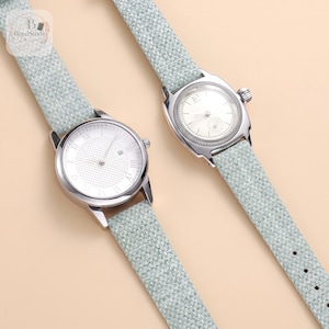 Peut inclure: Deux montres-bracelets avec des boîtiers en métal argenté et des bracelets en tissu vert clair texturé. La montre de gauche présente un cadran blanc avec des chiffres romains et un guichet de date. La montre de droite a un cadran blanc avec un seul chiffre à douze heures et une lunette cannelée.