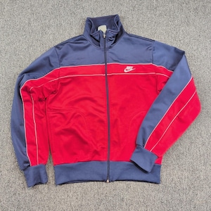 May include: A long-sleeve zip-up track jacket featuring a color-blocked design with a red torso and navy blue sleeves, shoulders, and collar. The jacket includes a white Nike logo on the upper right chest and thin white piping accents.