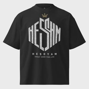 Op de afbeelding: Een zwart t-shirt met korte mouwen voorzien van een grote, grijze, getextureerde zeshoekige afbeelding met de letters HESOYAM. Boven de afbeelding staat een klein gouden kroonicoon. Onder de afbeelding staat de tekst HESOYAM en CHEAT CODE FOR LIFE in het wit.