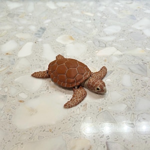 May include: A small, realistic figurine of a sea turtle with a textured, reddish-brown shell and patterned flippers. The turtle has a light-colored head with visible eyes and a small beak, captured in a detailed, lifelike pose with its flippers extended.