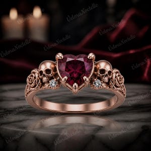 May include: A rose gold ring featuring a central heart-shaped deep red gemstone held by prongs. The band is detailed with two sculpted skulls, one on each side of the center stone, flanked by carved rose motifs and small round clear accent stones.