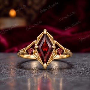 May include: A gold ring featuring a large, kite-shaped, deep red garnet gemstone at the center, flanked by two smaller, round, red garnet stones on either side. The metal band has an intricate, geometric, open-work design.