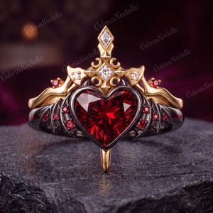 May include: A ring featuring a large heart-shaped red gemstone set in a black metal band with wing-like accents. The upper portion is crafted from gold-toned metal, adorned with small clear and red stones, and topped with a pointed, cross-like structure.