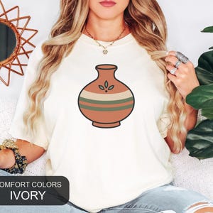 May include: An ivory t-shirt featuring a centered graphic of a terracotta-colored vase with horizontal stripes in cream and dark green, accented by a small three-leaf design. The shirt includes a black rectangular label reading COMFORT COLORS IVORY.