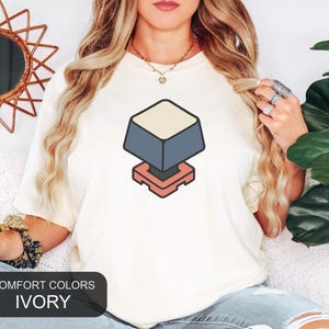 May include: An ivory t-shirt featuring a centered graphic of a mechanical keyboard keycap. The keycap is depicted in an isometric view with a cream-colored top, dark gray sides, and a coral-colored base. The shirt is labeled as Comfort Colors Ivory.