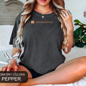 May include: A dark gray short-sleeve t-shirt with a crew neckline. The chest features a small orange graphic of a bus icon followed by the text "Hallucinating..." in a matching orange font. The fabric has a washed, vintage-style texture.