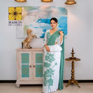 May include: A white two-piece outfit featuring a peplum top and a long skirt, both adorned with a large green lotus flower print. The ensemble includes a solid green shawl draped over one shoulder. The text MADE IN SRI LANKA is visible.