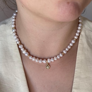 May include: A necklace featuring a strand of small, white, round pearls interspersed with tiny, polished gold-tone beads. A single, circular gold-tone pendant with a textured, hammered finish and a small, clear, central stone hangs from the centre.