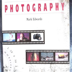 May include: A white hardcover book titled The Complete Encyclopedia of Photography by Mark Edwards. The cover features two horizontal film strips containing six small photographs and a circular aperture scale graphic with numbers 2 through 16.