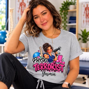 May include: A heather gray short-sleeve t-shirt featuring a graphic illustration of a person in blue scrubs holding a massage gun, surrounded by a blue exercise ball, a spine model, and a pink foam roller, with the text "PHYSIO TERRORIST Yasmin".