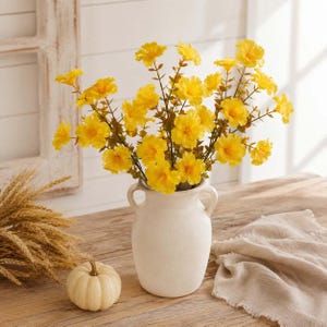 May include: A bouquet of bright yellow flowers with thin brown stems arranged in a matte white ceramic vase with two side handles. The flowers feature five rounded petals each and are displayed in a full, rounded cluster with green foliage.