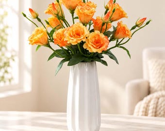 May include: A bouquet of vibrant orange eustoma flowers with green stems and leaves arranged in a tall, white, faceted ceramic vase. The flowers feature multiple layers of delicate petals, with some buds still closed, creating a full, rounded display.