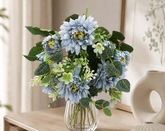 May include: A bouquet of artificial light blue chrysanthemum-like flowers with yellow centers, accented by small pale green blossoms and dark green leaves, arranged in a clear, vertically ribbed glass vase with a rounded base.