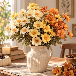 May include: A ceramic pitcher with a cream-colored speckled finish and vertical ribbed texture holds a large bouquet of artificial flowers in shades of white, yellow, and burnt orange, featuring green leaves and prominent yellow centers.