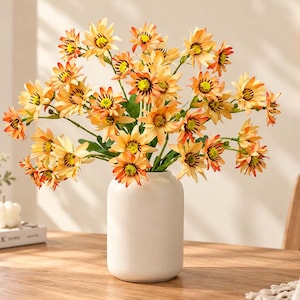 May include: A bouquet of artificial orange and yellow daisy-like flowers with dark centers and green stems arranged in a smooth, matte white cylindrical vase. The petals feature gradient coloring, transitioning from light yellow tips to orange bases.