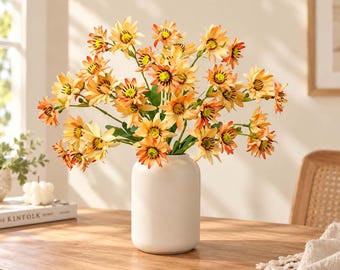 May include: A bouquet of artificial orange and yellow daisy-like flowers with dark centers and green stems arranged in a smooth, matte white cylindrical vase. The petals feature gradient coloring, transitioning from light yellow tips to orange bases.