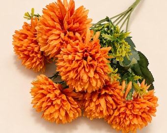 May include: A bouquet of artificial orange chrysanthemum flowers with layered petals. The arrangement features multiple round blooms attached to green stems with serrated green leaves and small sprigs of foliage extending from the base.