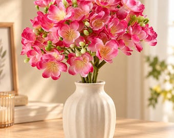 May include: A bouquet of vibrant pink and white artificial flowers with small green buds arranged in a cream-colored, vertically ribbed ceramic vase. The flowers feature five petals each with yellow centers, creating a dense, rounded floral display.
